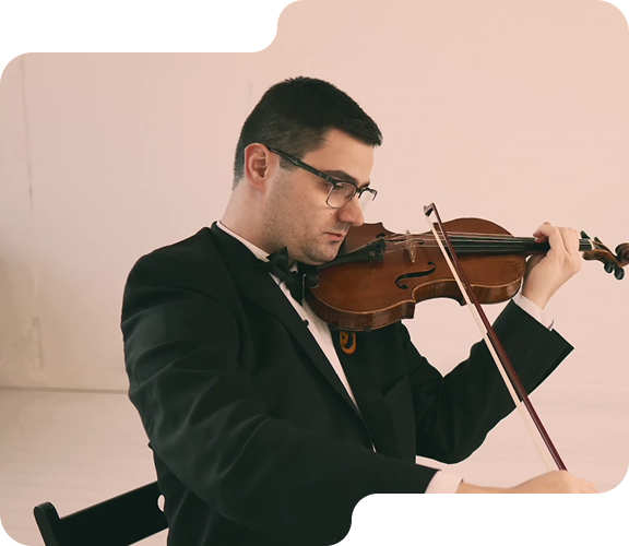 Man in tuxedo playing violin with focused expression, seated on a black chair against a plain background.