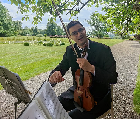 Man in black suit with glasses playing violin outdoors near a music stand and garden path.