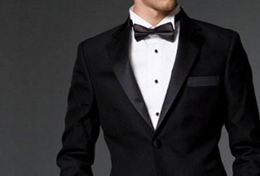 Close-up of man wearing black tuxedo jacket, white shirt, and black bow tie against dark background.